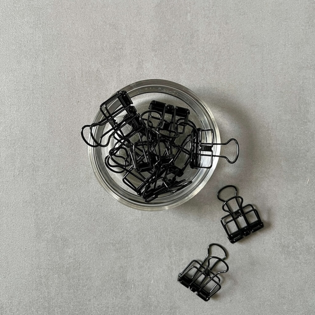 Black binder clips on a light gray surface with a clear dish.