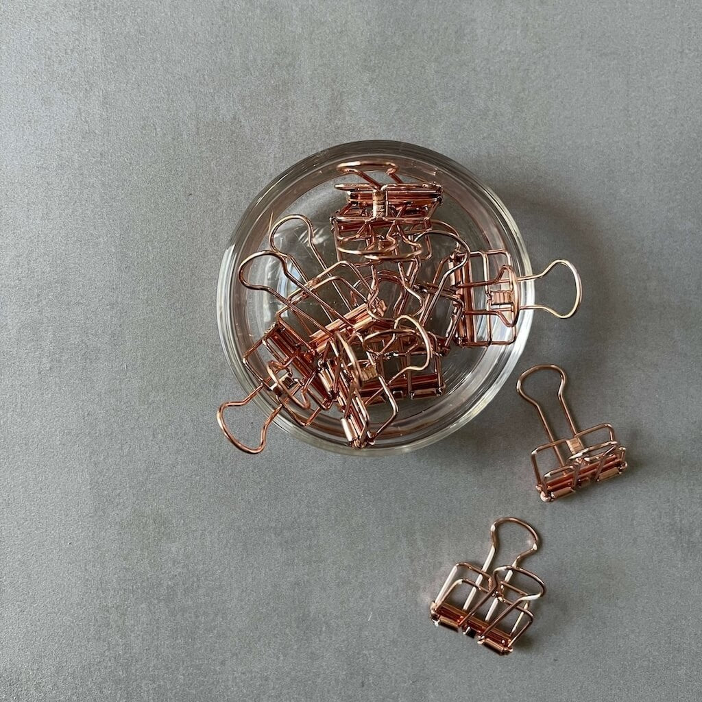 Rose gold binder clips on a gray surface with a clear glass dish.